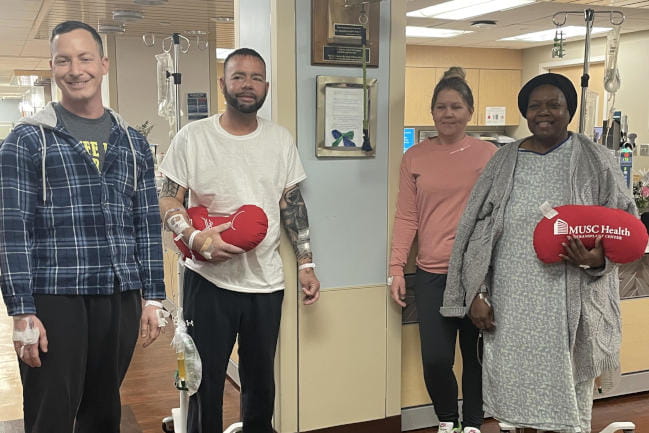 Four people stand in a hospital waiting room, some with IVs attached. They are holding plush pillows shaped like kidneys with the 沙巴体育金刚官网入口 Health logo printed on them.